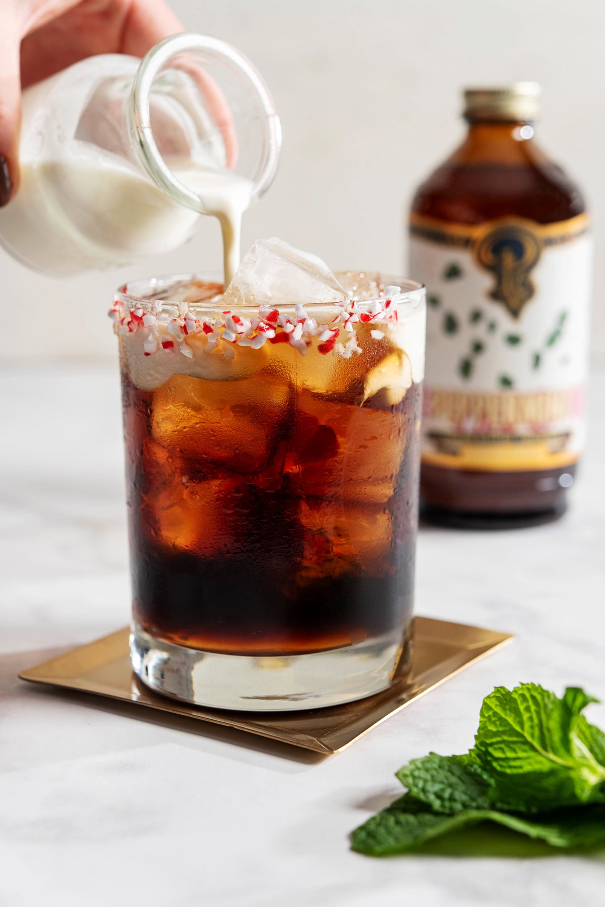 Pouring milk into a glass of iced coffee with peppermint candy on a white background