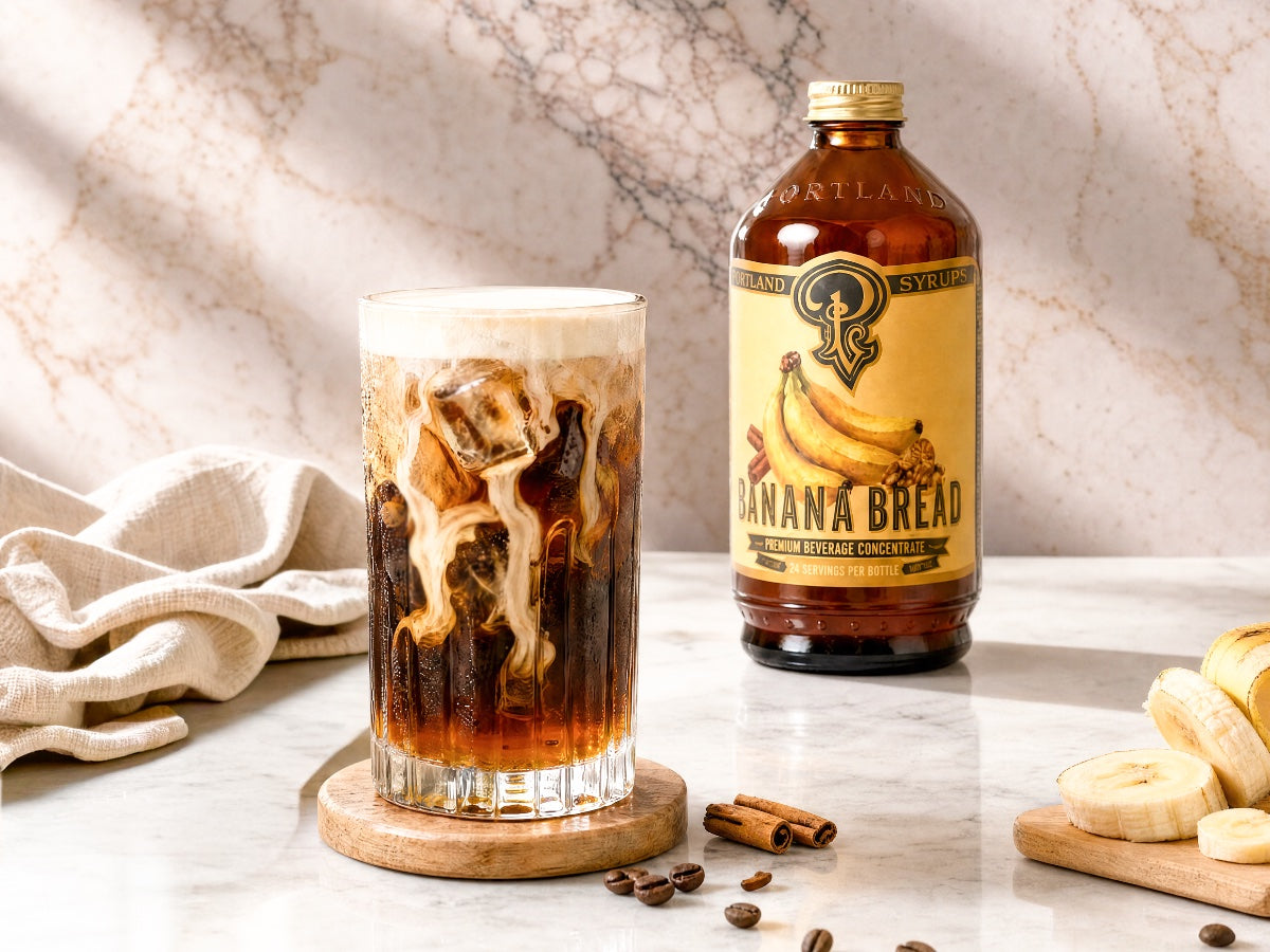 Banana Bread-flavored syrup bottle with a glass of iced coffee on a marble surface.