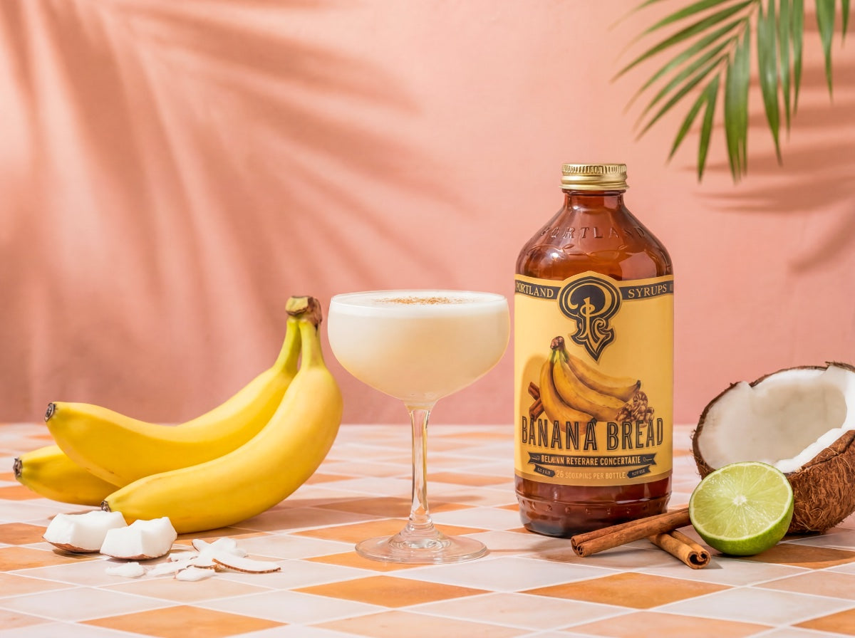 Banana Bread syrup bottle with a glass of a creamy drink, bananas, coconut, lime, and cinnamon sticks on a checkered tablecloth.