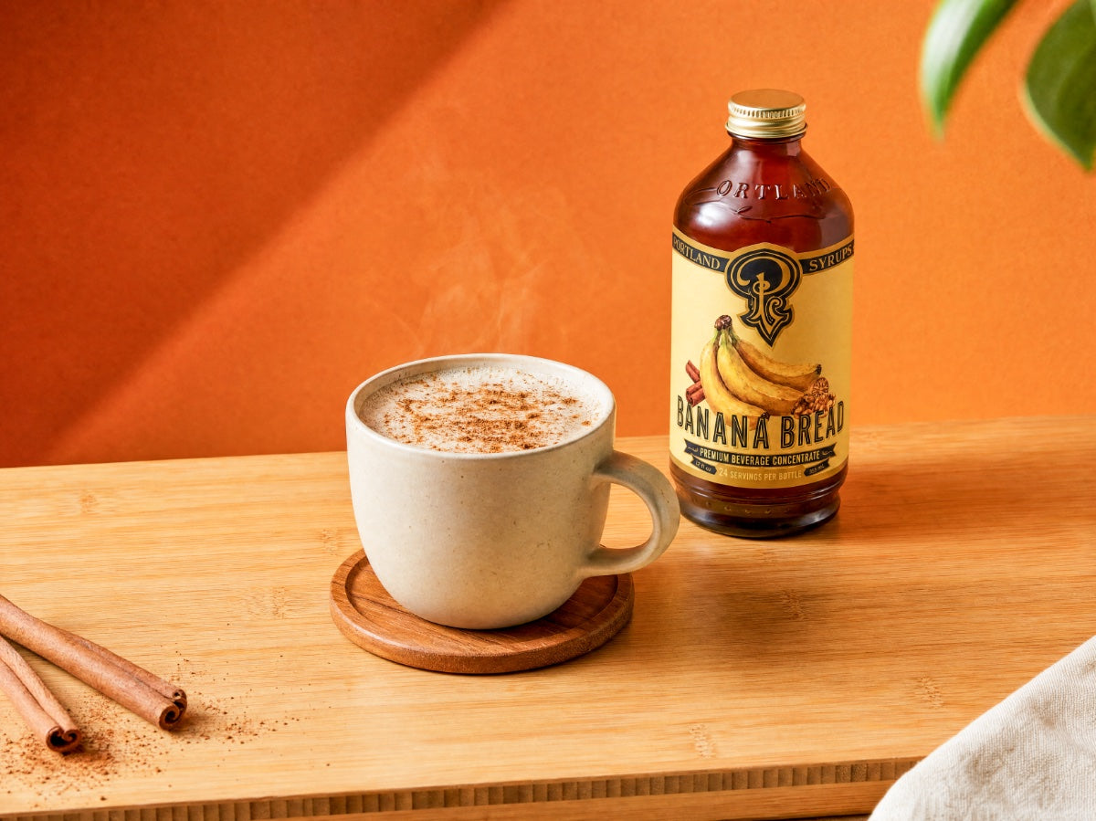Banana bread-flavored coffee bottle next to a steaming cup of coffee on a wooden surface with an orange background.