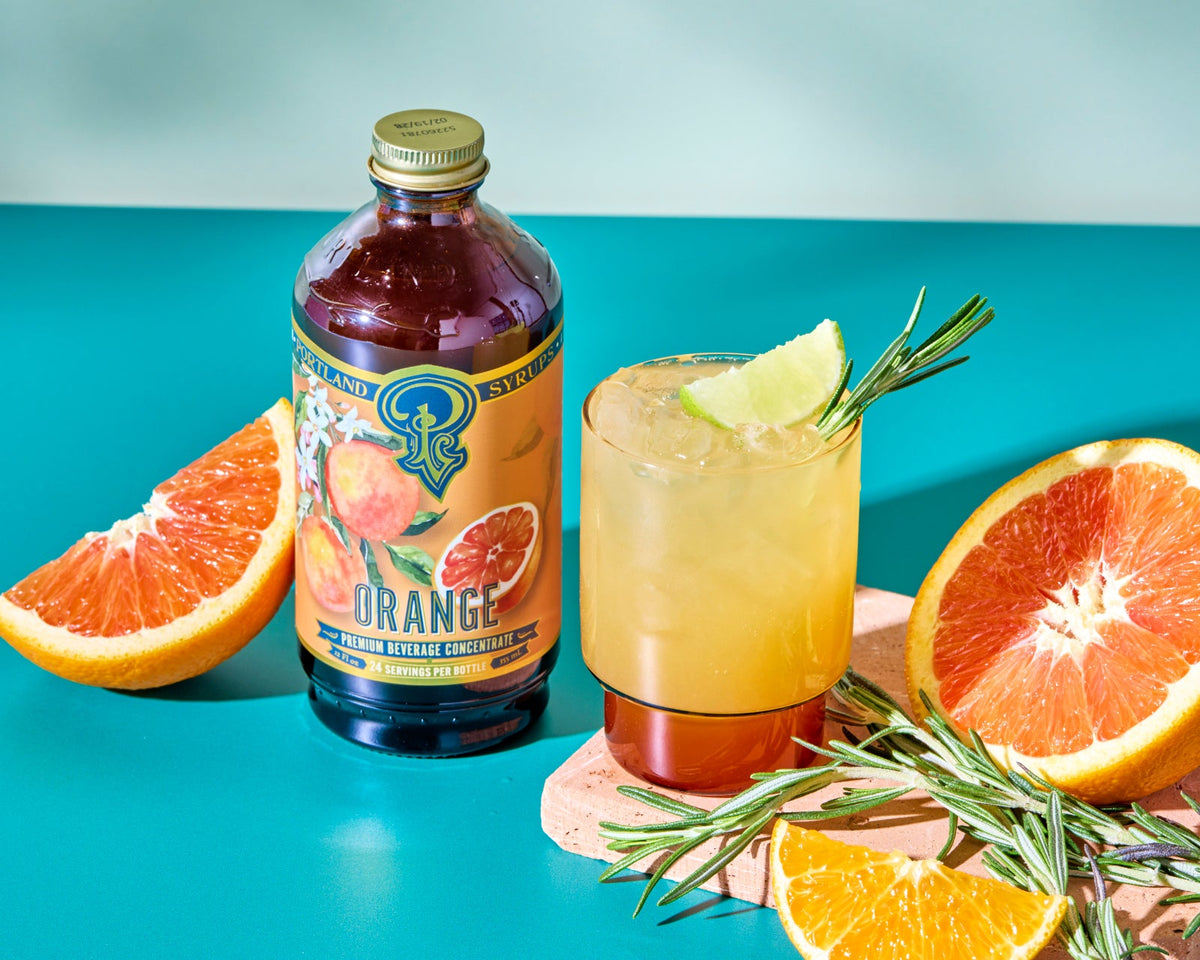 Bottle of orange juice concentrate with a glass of orange juice, lime, and rosemary on a teal background