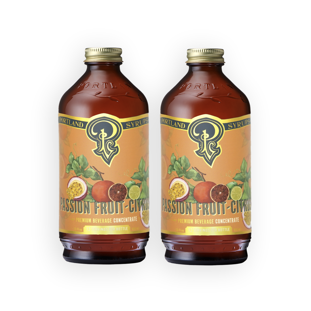 Two amber glass bottles of Portland Syrup Passion Fruit Citrus beverage concentrate with gold caps and orange labels, shown side by side on a white background.