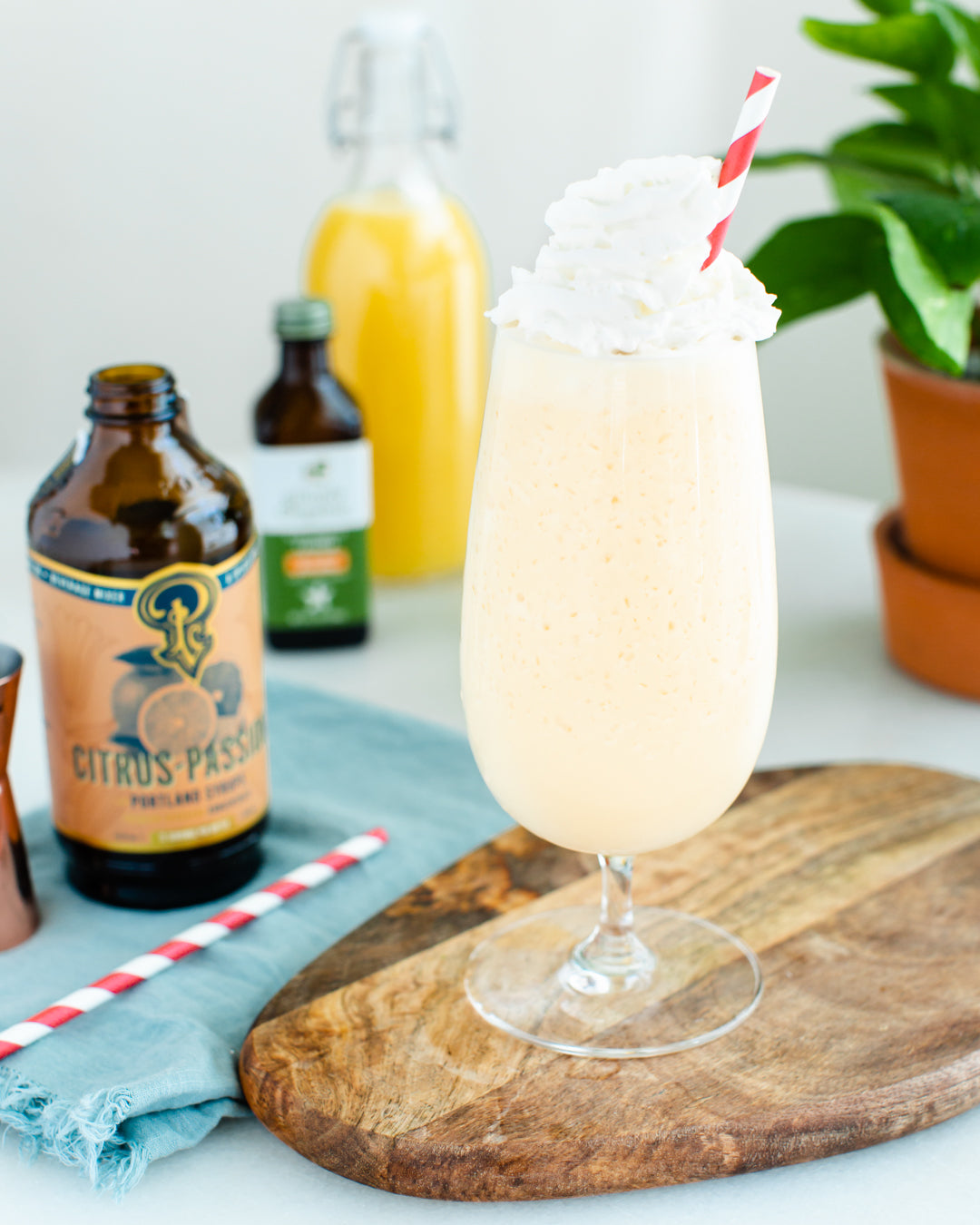 Glass of creamy drink with whipped cream and a straw on a wooden board, with bottles and a plant in the background.