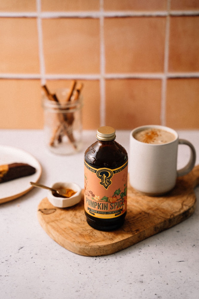 Bottle of pumpkin spice syrup on a wooden cutting board with a cup of coffee and a plate of food in the background.