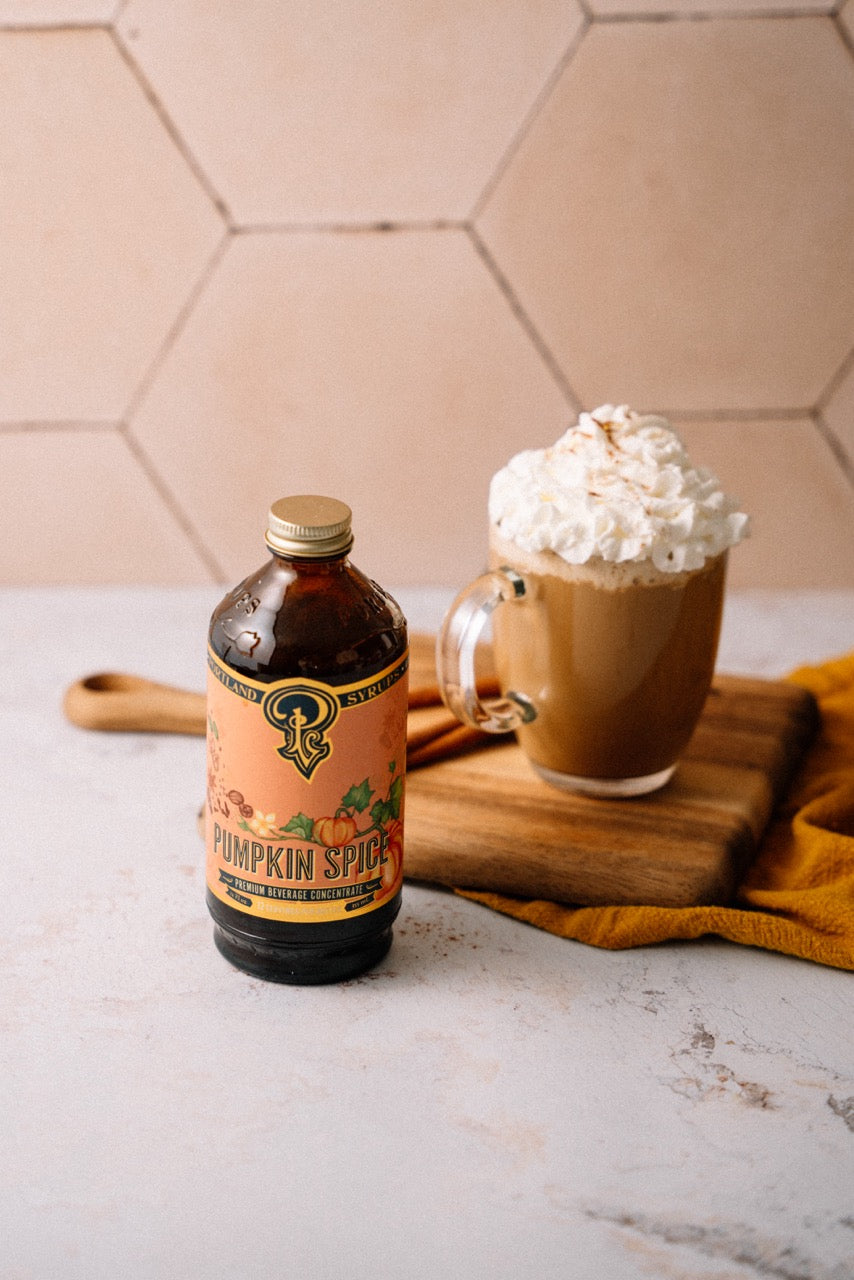 Bottle of pumpkin spice syrup next to a mug of hot chocolate with whipped cream on a hexagonal tile background.
