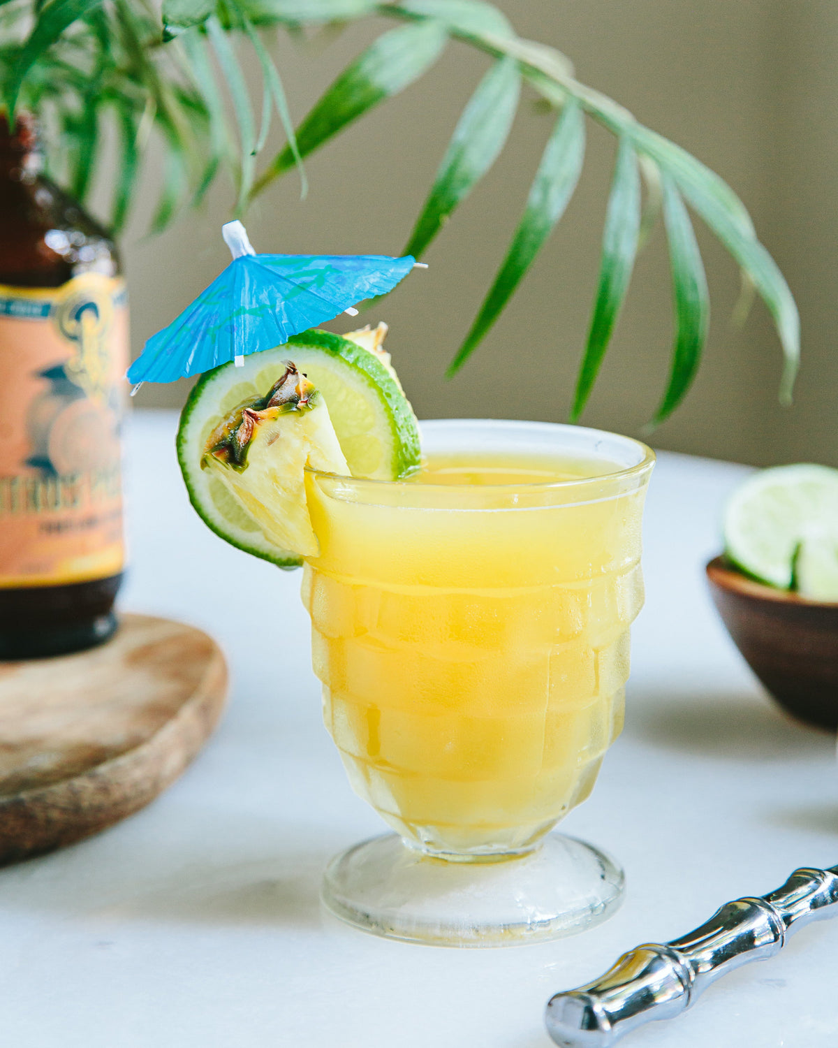 Glass of yellow cocktail with lime and blue umbrella on a white surface with a plant in the background