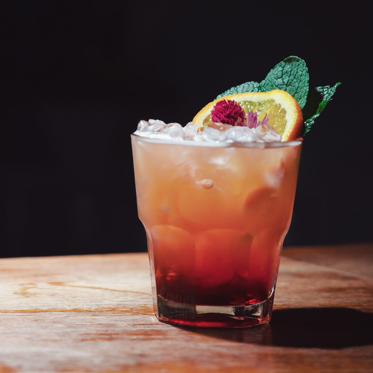 Cocktail with ice, garnished with an orange slice and mint leaf on a wooden surface.