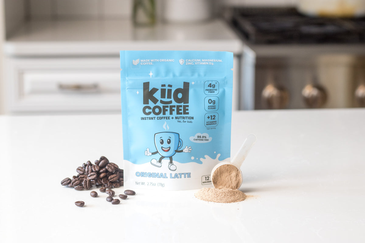 A stand-up pouch of kiid COFFEE Organic Latte powder sits on a white countertop with coffee beans and a spoonful of the light brown powder in front of a blurred kitchen background.