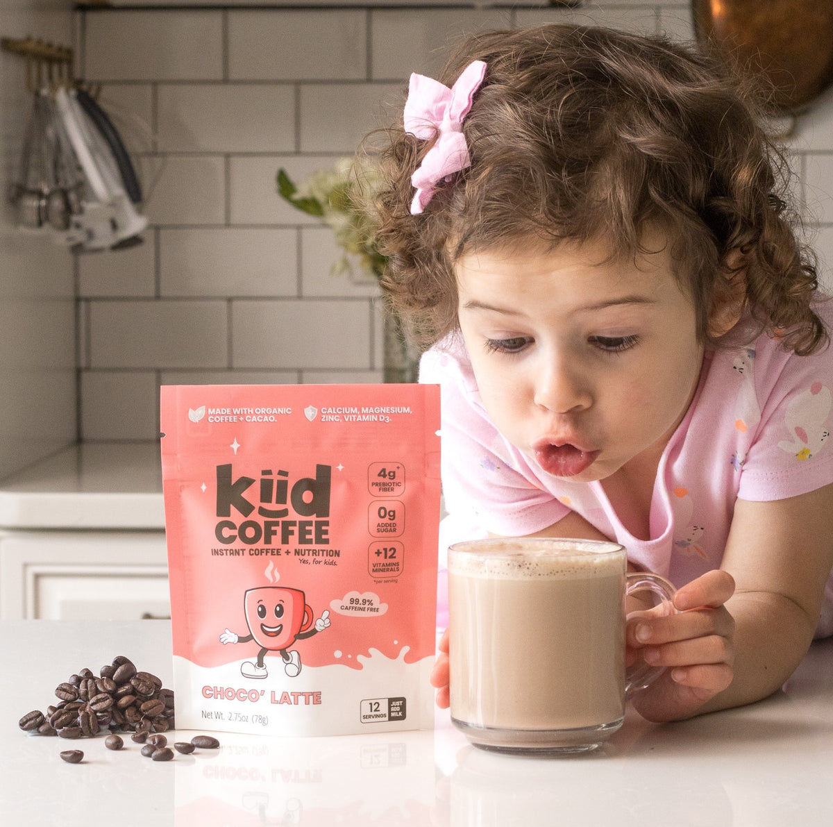 A young girl with curly brown hair and a pink bow leans over a counter, drinking from a glass mug of coffee latte. Beside her is a pink pouch of kiid COFFEE Choclo Latte, with coffee beans scattered on the counter.
