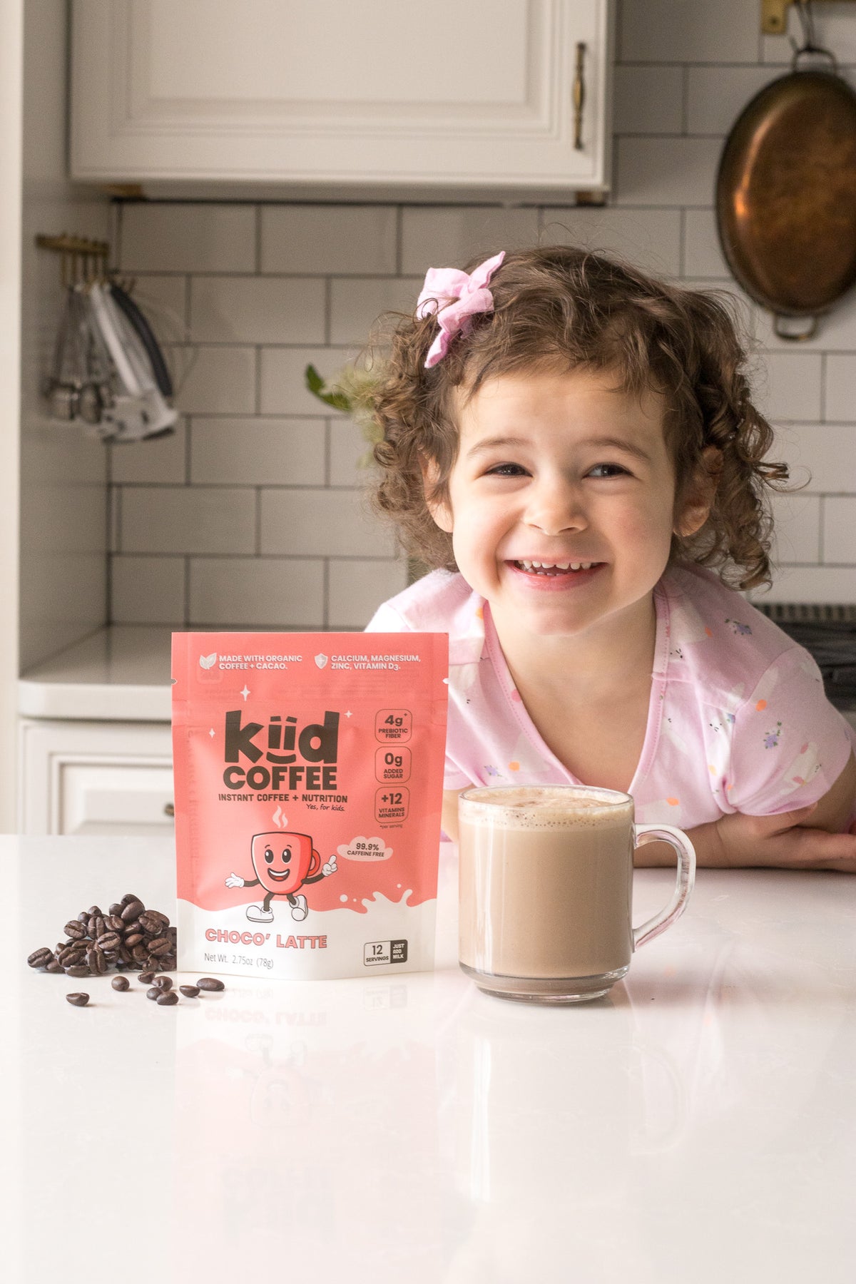 A young girl with curly brown hair and a pink bow smiles behind a counter, with a bag of Kid Coffee Brood Latte and a glass of the prepared drink in a bright kitchen setting.