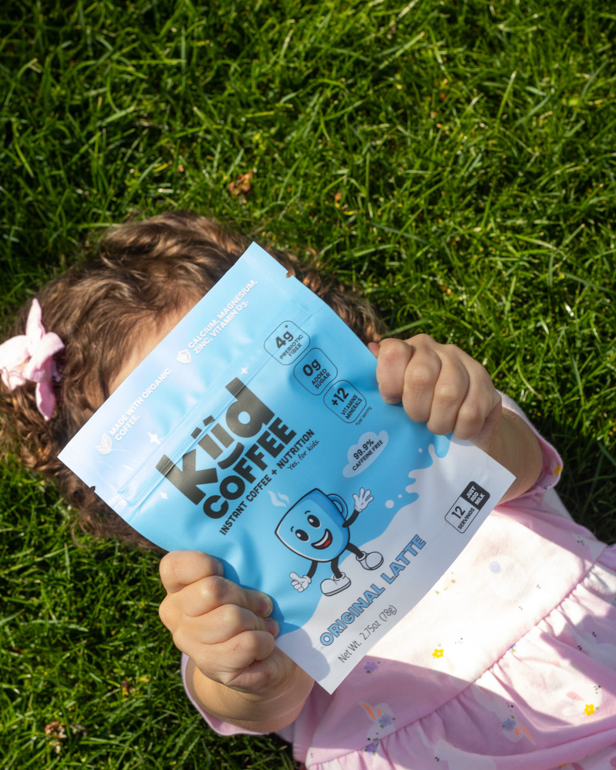A young child lying on green grass holds a light blue bag of Kiid Coffee Kid Coffee + Protein Original Latte over their face. The child is wearing a light pink dress and has a pink bow in their hair.