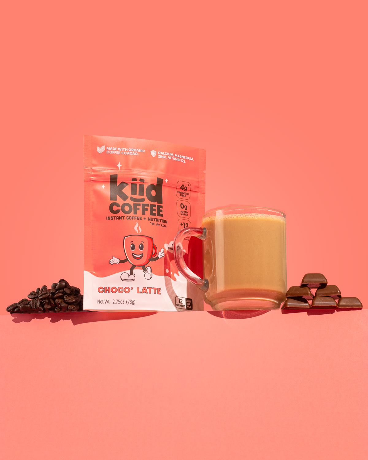 A pouch of kiid COFFEE Choco Latti instant coffee next to a glass mug filled with a choco latte, styled with chocolate chips and chocolate pieces on a solid coral pink background.