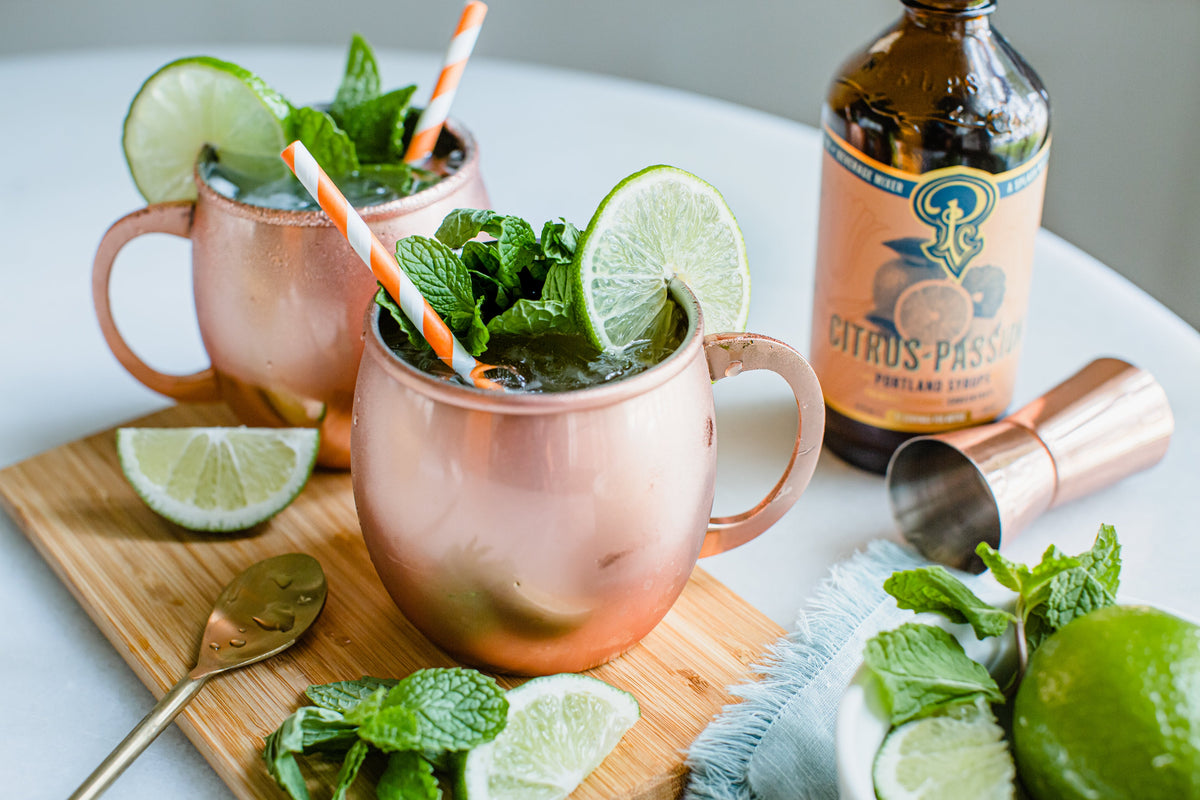 Two copper mugs with mint and lime cocktails on a wooden board, accompanied by a bottle of Citrus Passport and bar tools.
