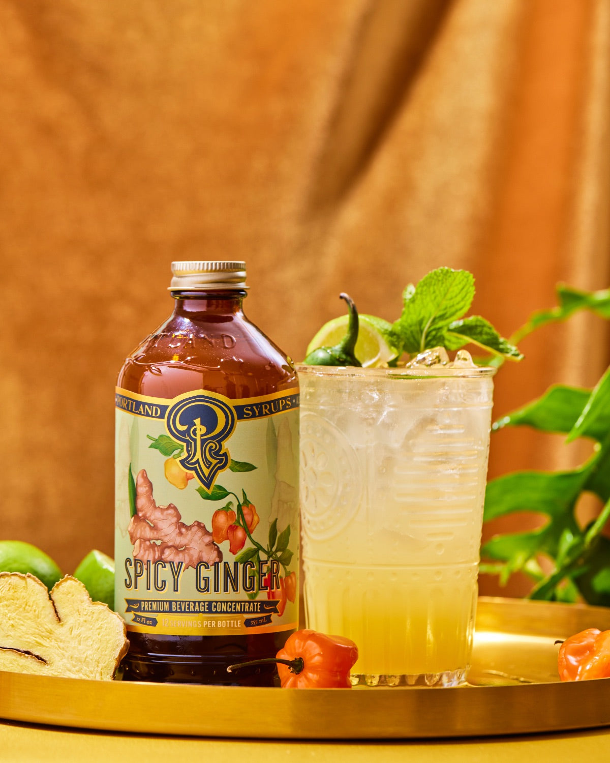 A bottle of Spicy Ginger hot pepper extract next to a prepared cocktail in a clear glass, garnished with lime, mint, and ice, set on a gold tray with tropical ingredients against a warm brown backdrop.