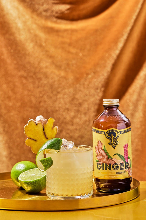 A bottle of ginger beer with a yellow label sits on a gold tray next to a glass of pale yellow cocktail with ice, garnished with a lime wedge, a lime half, and a ginger root, set against a draped gold fabric background.
