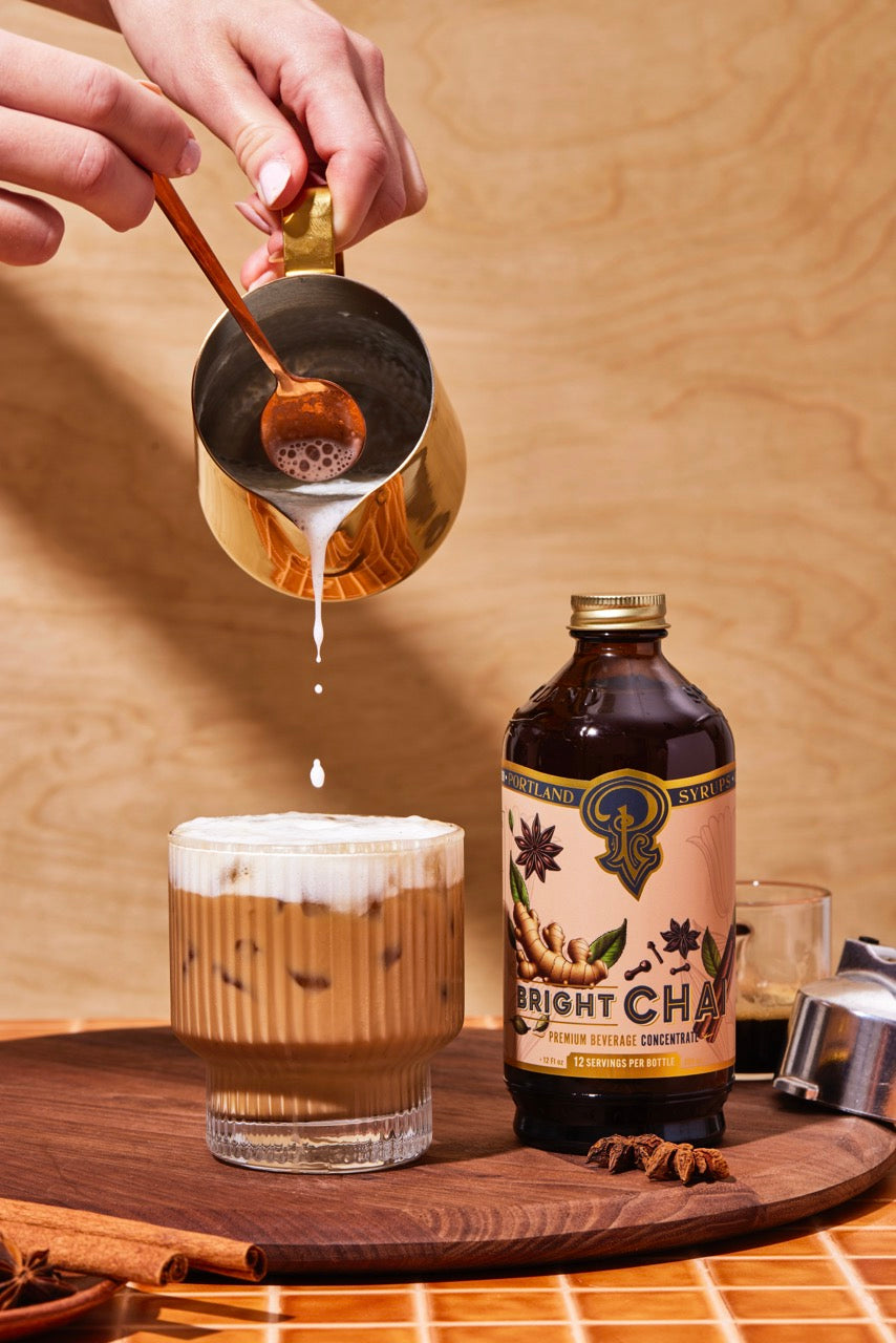 Person pouring a drink from a gold pitcher into a glass with a bottle of 'Bright Chai' in the background.