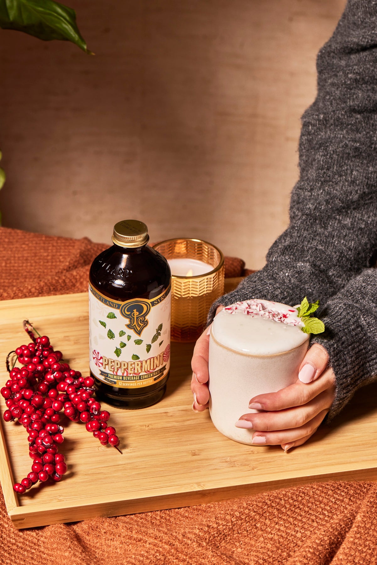 Person holding a mug of peppermint hot chocolate with a bottle of peppermint extract on a wooden tray.