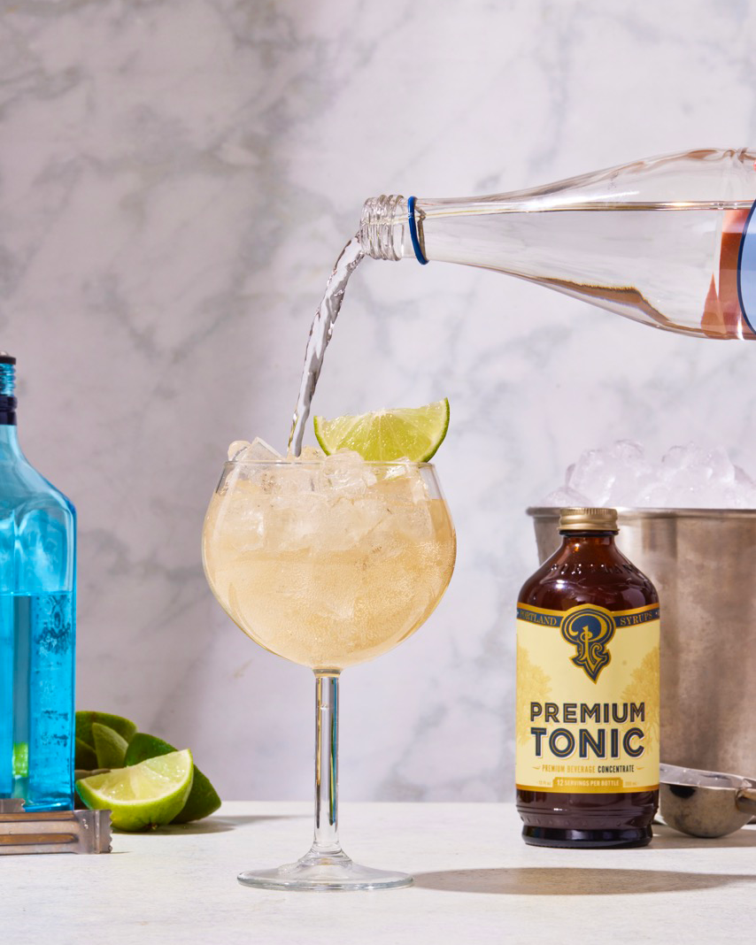 Pouring tonic into a cocktail with lime wedges, surrounded by bottles on a marble surface.