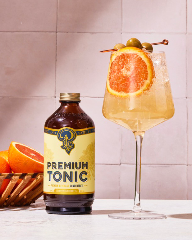 Bottle of Premium Tonic with a cocktail garnished with an orange slice and olive on a tiled wall background.