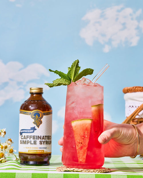 Caffeinated simple syrup bottle with a pink cocktail garnished with watermelon and mint leaves against a blue sky background.