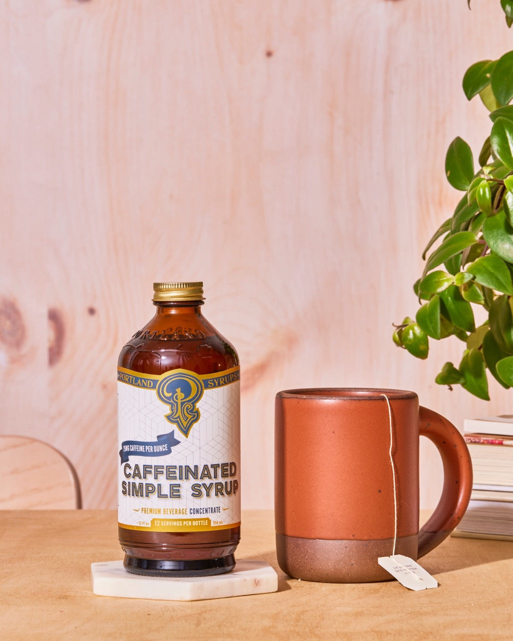 Bottle of 'Caffeinated Simple Syrup' next to a terracotta mug on a wooden surface with a plant in the background.