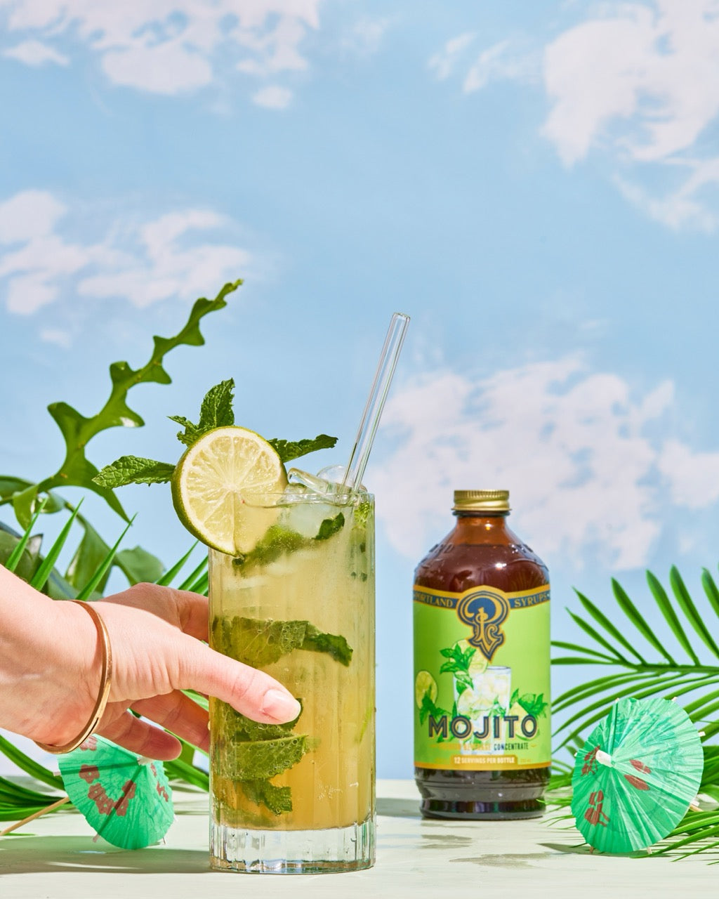 Mojito cocktail with lime and mint leaves, bottle of mojito mix, and palm leaves against a blue sky.