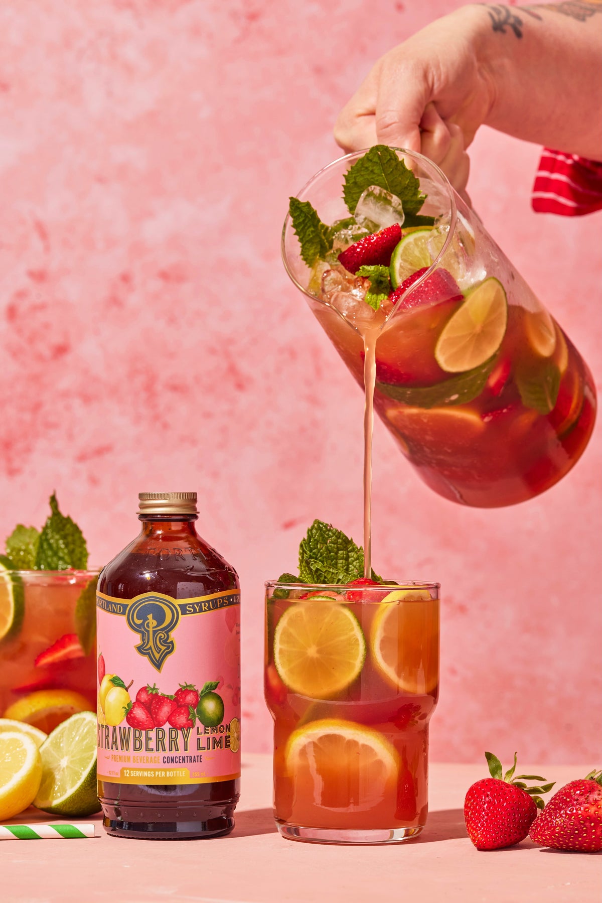 Strawberry lime iced tea being poured into a glass with fresh fruits and mint leaves on a pink background.