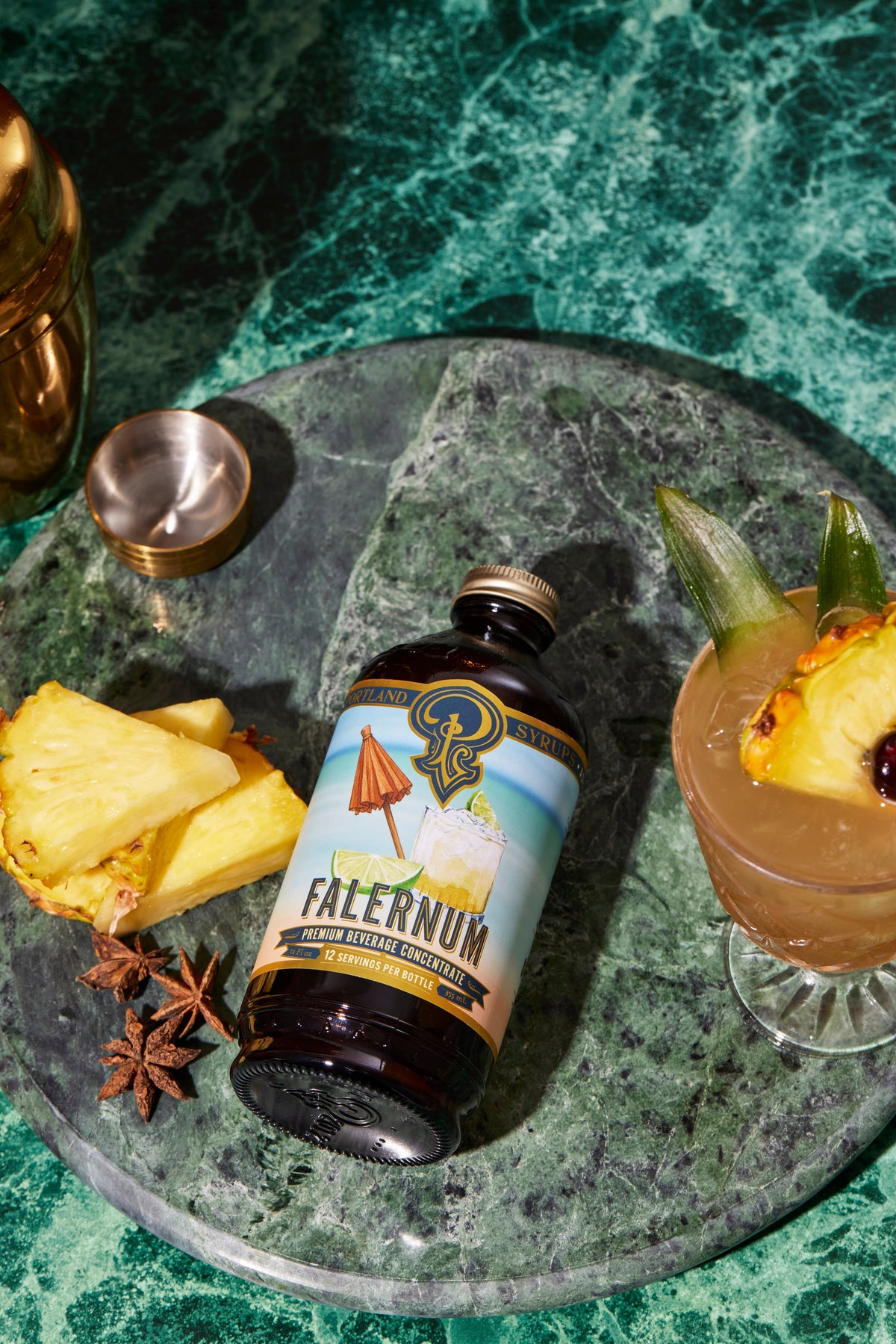 Bottle of Falernum syrup on a green marble surface with a cocktail and pineapple garnish.
