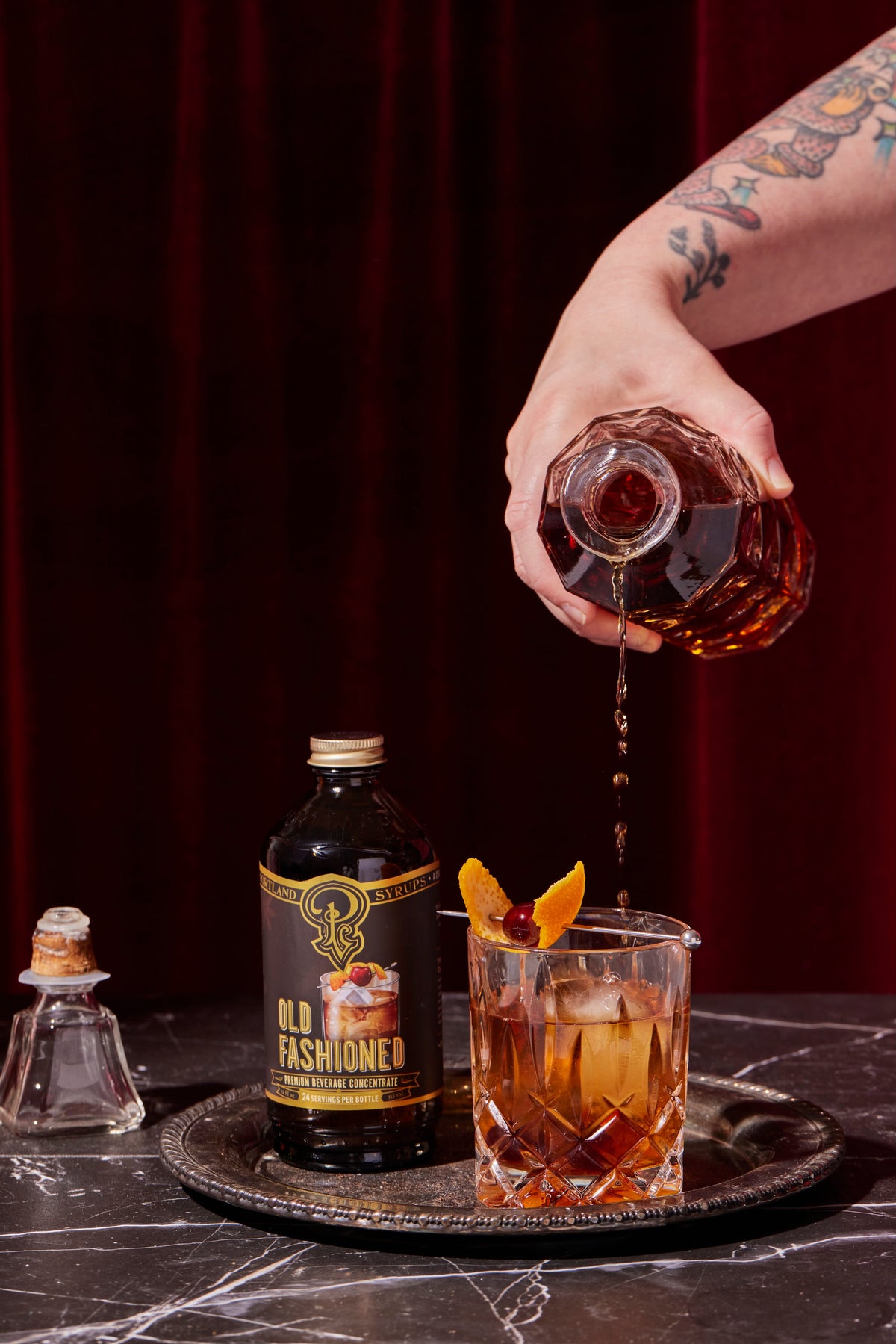 Hand pouring Old Fashioned cocktail into a glass with a bottle of Old Fashioned mix on a dark surface.