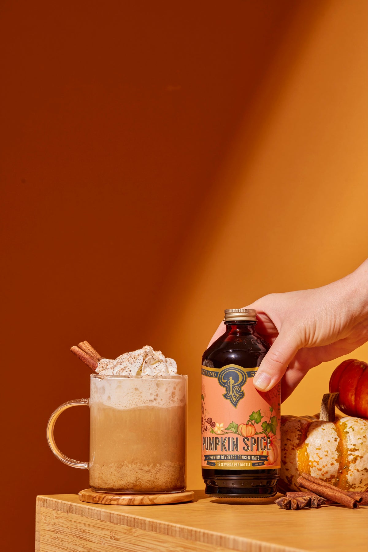 Hand holding a bottle of pumpkin spice syrup next to a mug of pumpkin spice latte on an orange background