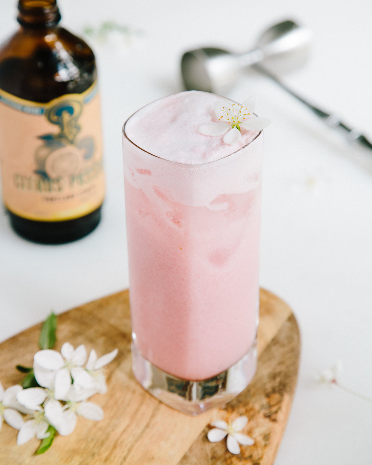 Pink cocktail in a glass with ice and flowers, next to a bottle and utensils on a white surface.