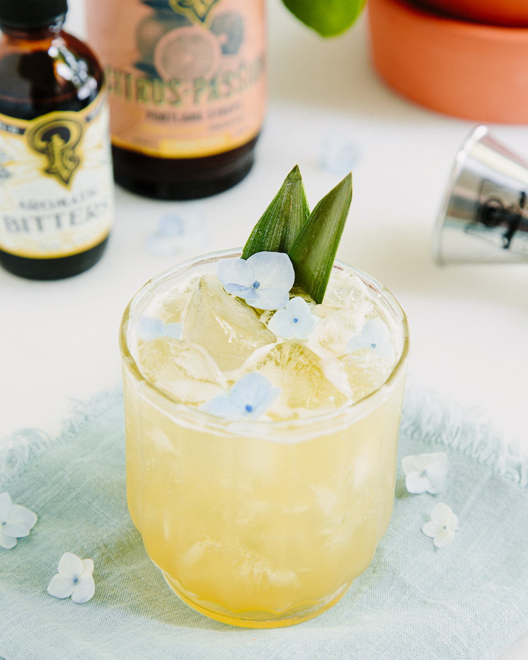Cocktail with floral garnish in a glass, surrounded by bottles and flowers on a light surface.