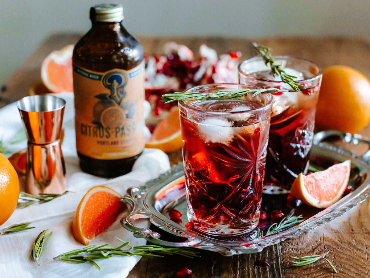 Two glasses of red cocktails with ice and garnishes on a tray, accompanied by a bottle of Citrus Paso and citrus fruits.