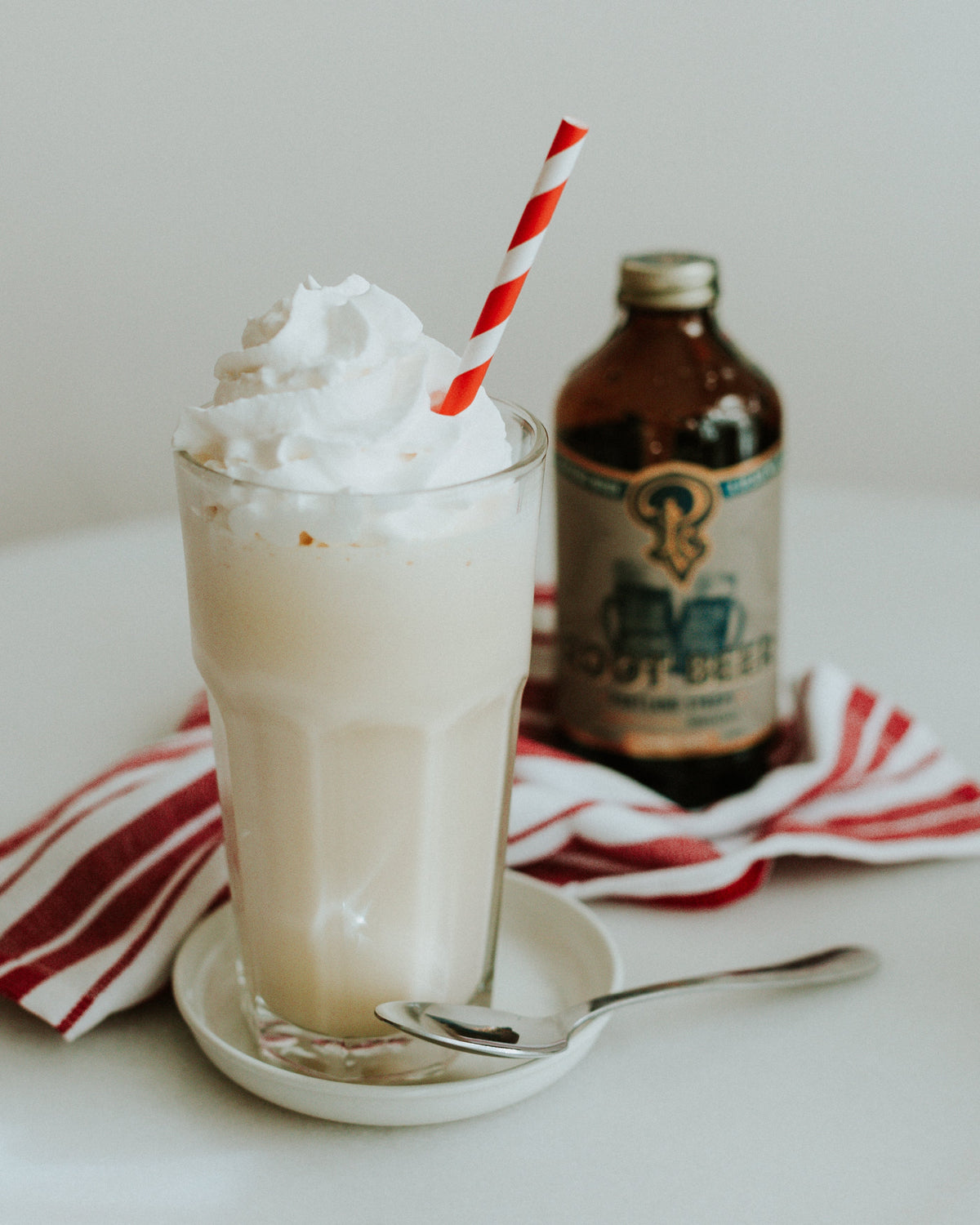 Glass of iced coffee with whipped cream and a red and white straw, next to a bottle of coffee on a striped cloth.