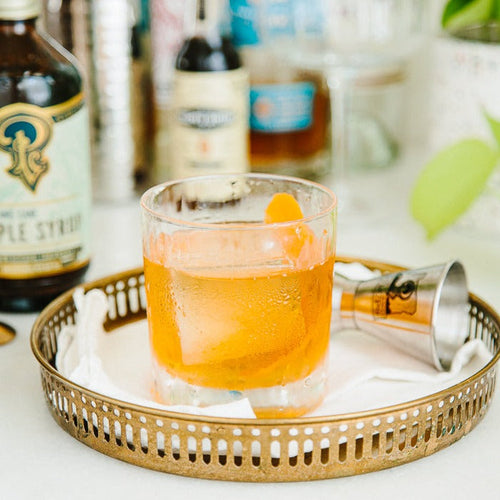 Cocktail in a glass with ice and an orange slice on a gold tray, surrounded by bottles.