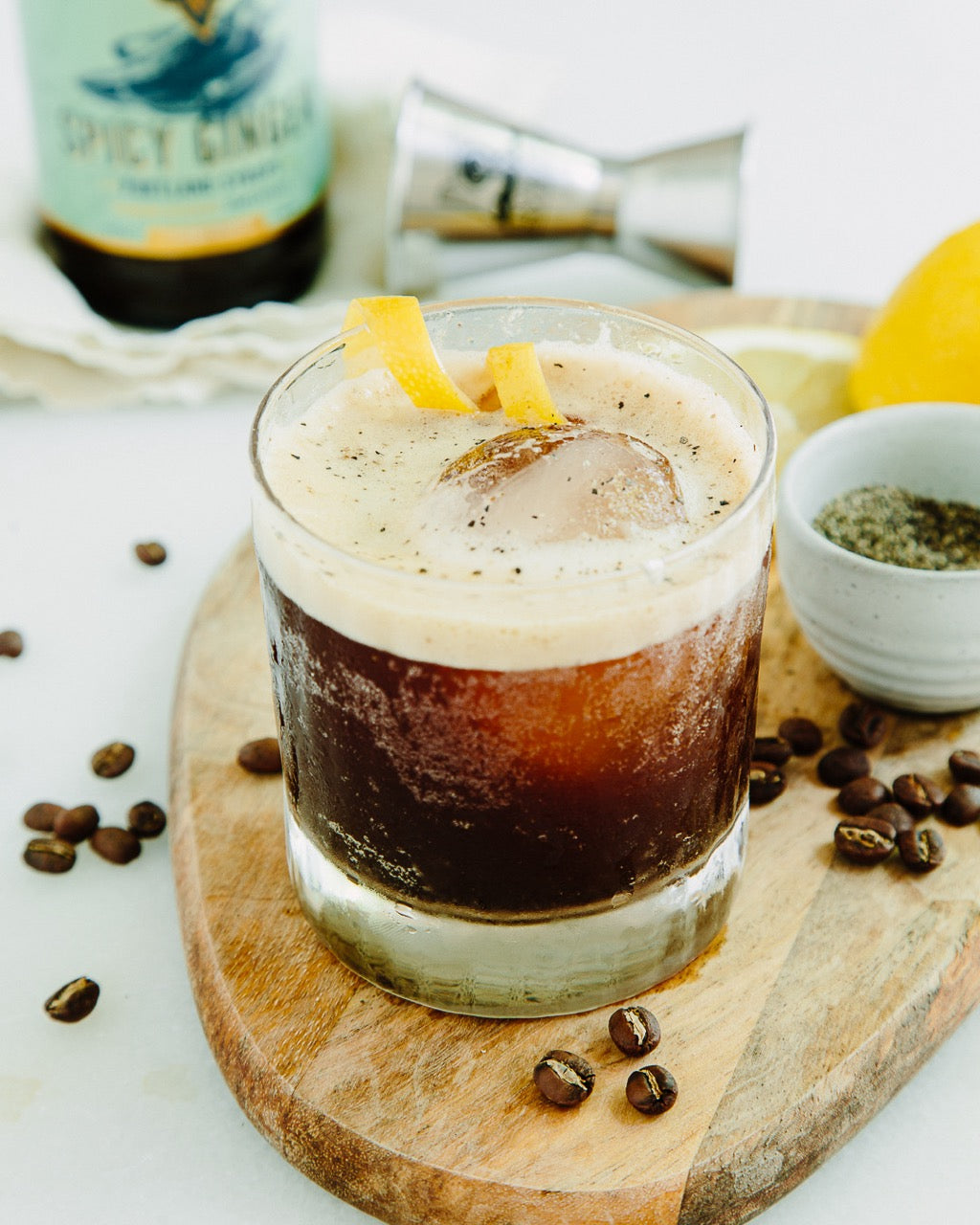 Glass of coffee cocktail with lemon peel on a wooden board with coffee beans and a bottle in the background.