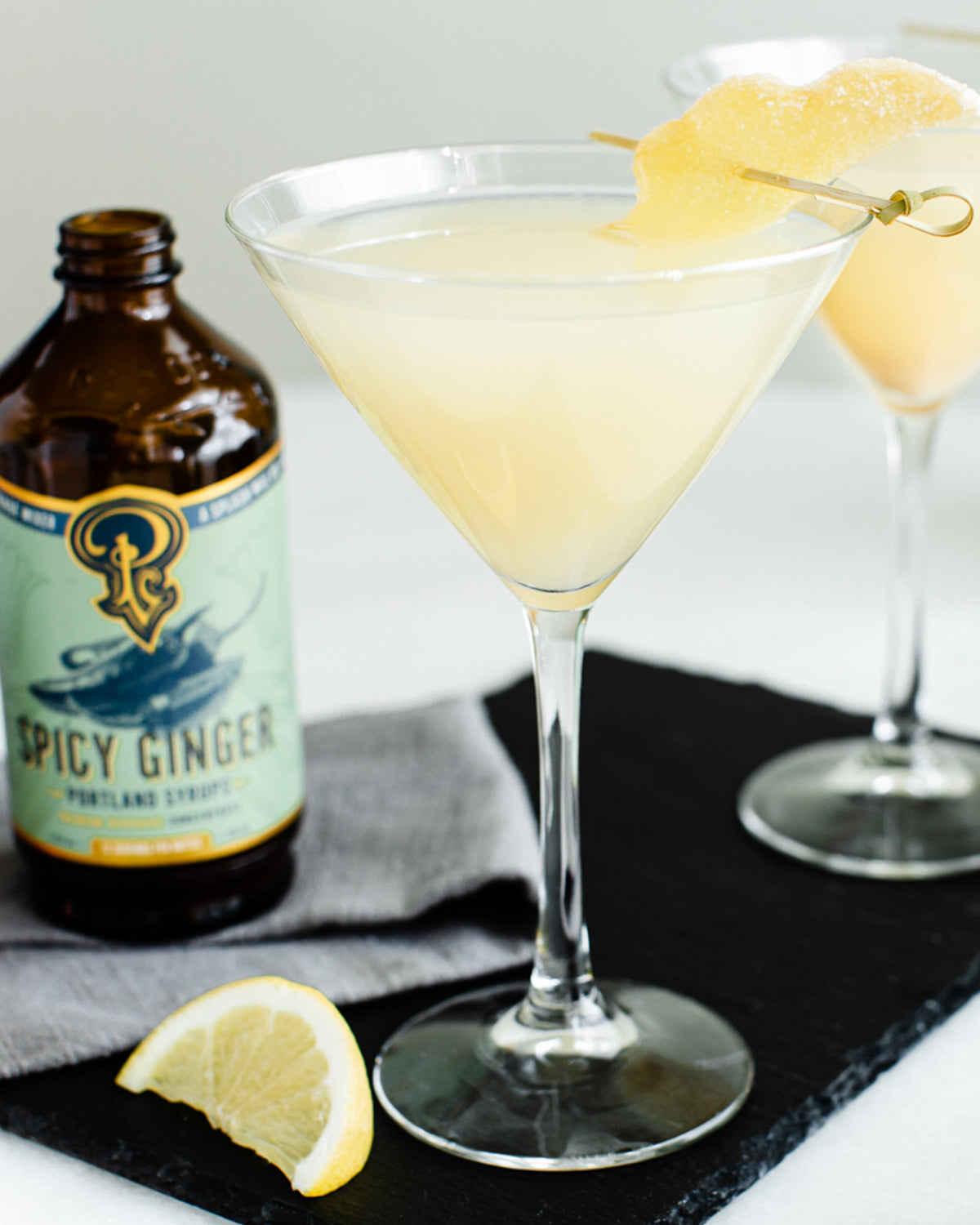 Cocktail in a martini glass with a bottle of Spicy Ginger on a dark slate surface.