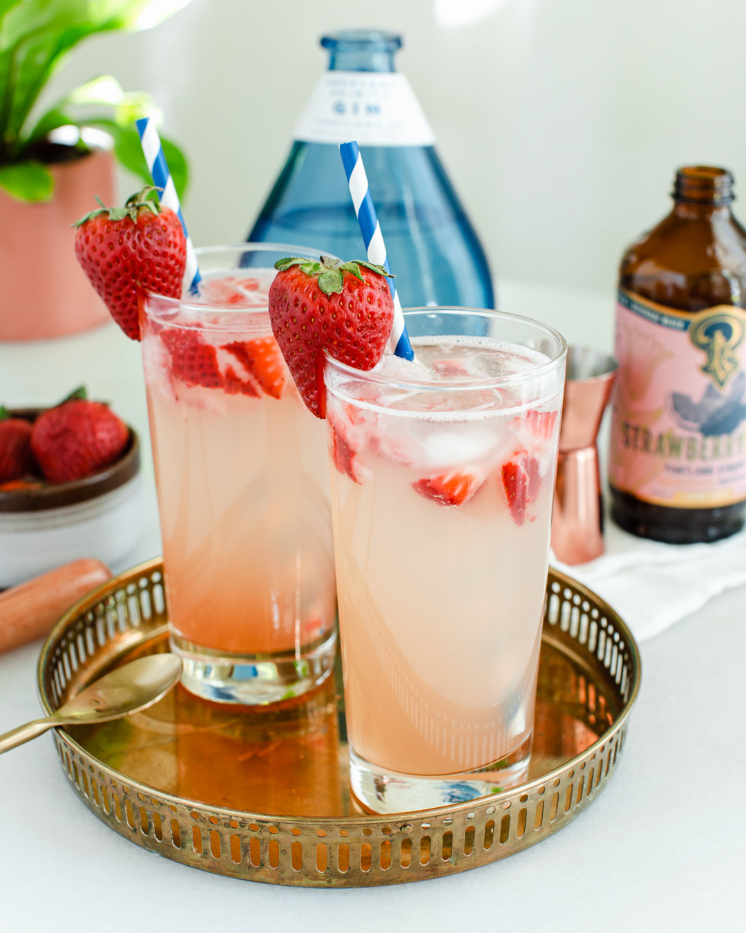 Two glasses of strawberry lemonade with ice and strawberries on a gold tray, with a bottle of strawberry lemonade in the background.