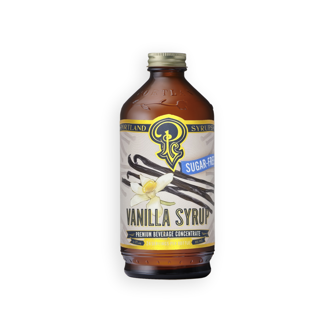 An amber glass bottle of Vanilla Syrup with a cream-colored label, featuring vanilla bean illustrations and a gold cap, set against a plain white background.