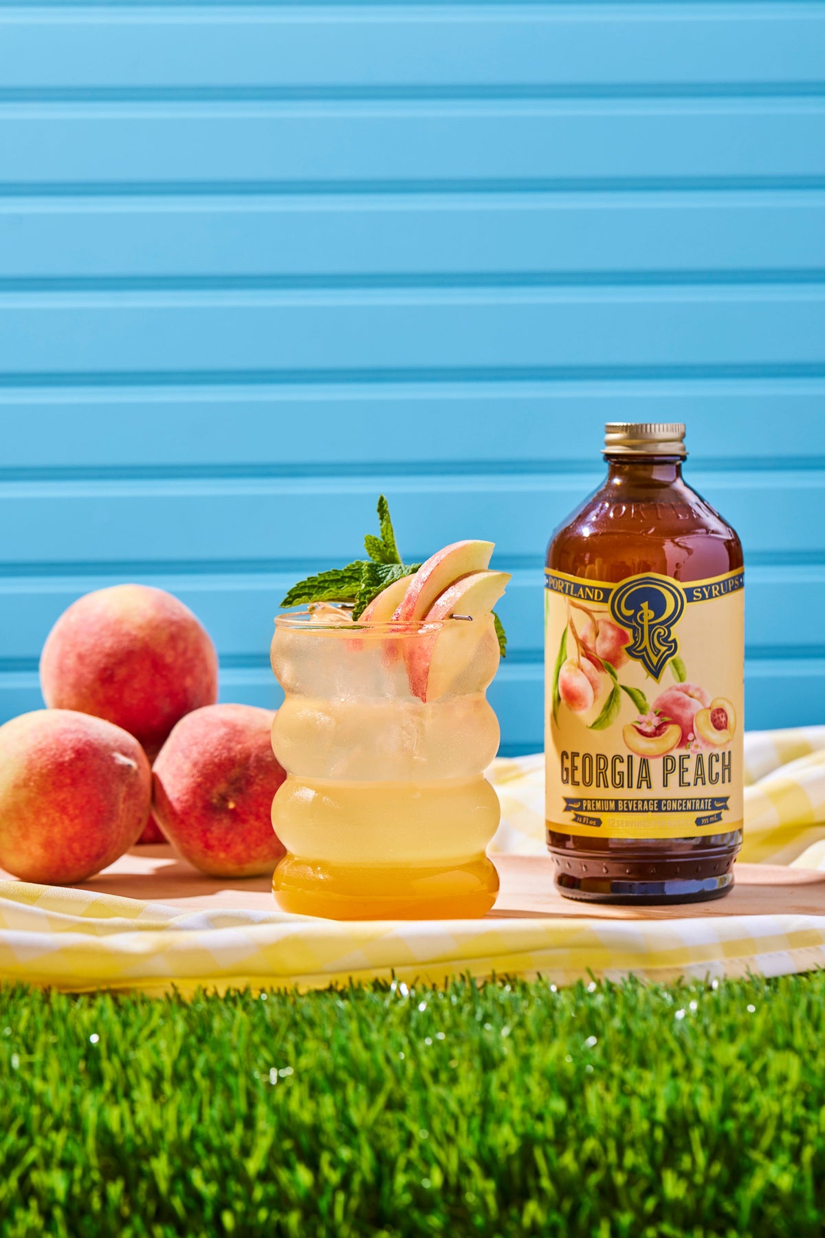 Georgia Peach cocktail with a bottle of Georgia Peach syrup on a grassy area with peaches and a blue background.